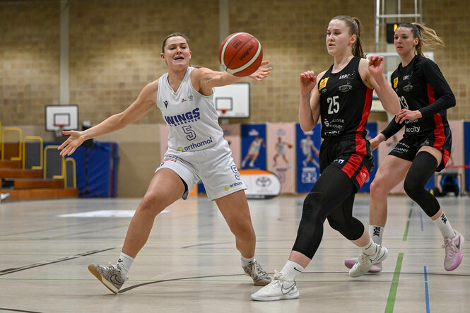 Basketballspielerin streckt die Hand seitlich aus, um Ball zu erreichen, zwei Gegenspielerinnen im Hintergrund