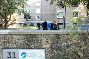 Eine Gruppe von Menschen steht vor einem mehrstöckigen Gebäude. Im Vordergrund ist eine Mauer mit einem Schild, auf dem "31" und das Logo sowie der Name "European Homecare" zu sehen sind. Bäume und Pflanzen rahmen die Szene ein.