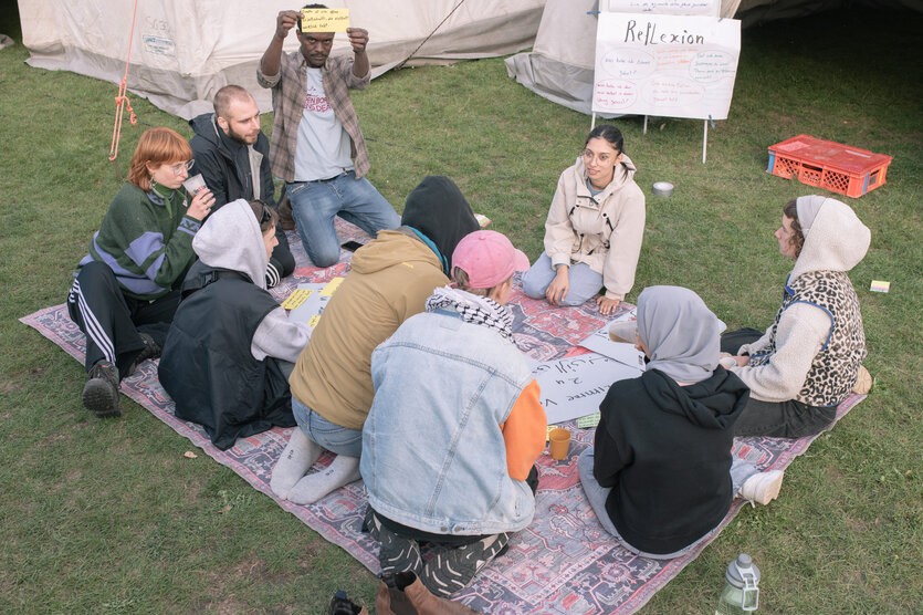 Eine Gruppe von Menschen sitzt auf einer Wiese im Kreis. Im Hintergrund steht auf einer Tafel das Wort "Reflexion"