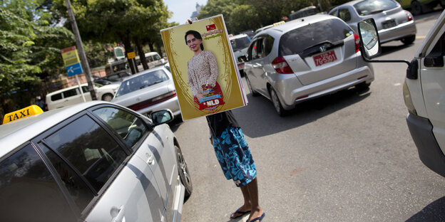 Ein Mann steht zwischen Autos auf der Straße in Birma. Er hält ein Plakat mit Aung San Suu Kyi, es verdeckt seinen Rücken und das Gesicht Ein Mann steht zwischen Autos auf der Straße in Birma. Er hält ein Plakat mit Aung San Suu Kyi, es verdeckt seinen Rücken und das Gesicht