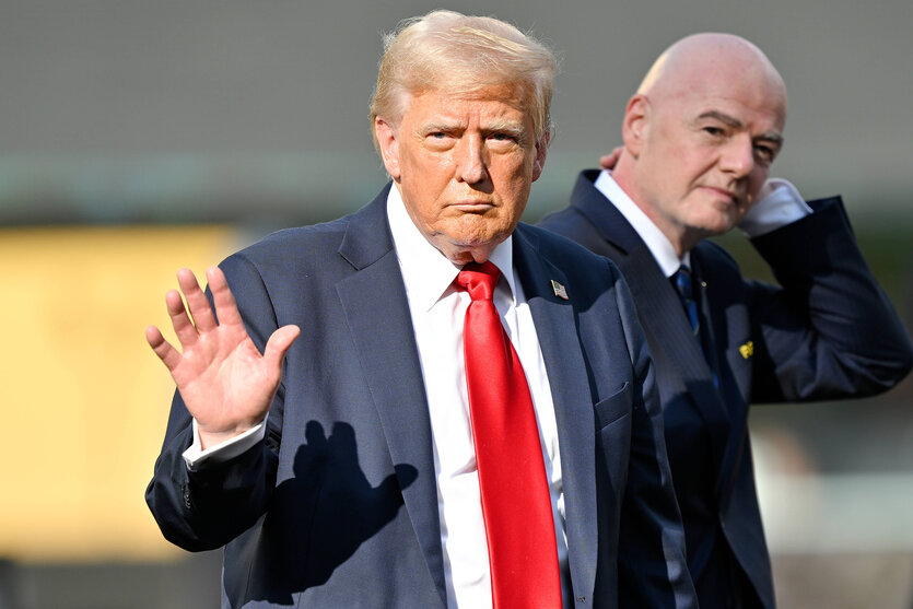 Donald Trump winkt, dahinter steht Gianni Infantino und beobachtet ihn