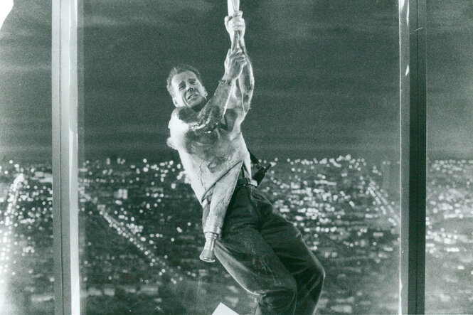 Bruce Willis in "Stirb Langsam". Er baumelt an einem Seil vor einem Fenster. Hinter ihm ist die Stadt von oben sichtbar. Er befindet sich also sehr hoch oben.