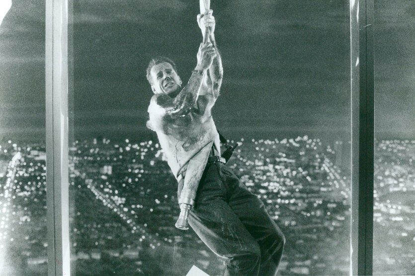 Bruce Willis in "Stirb Langsam". Er baumelt an einem Seil vor einem Fenster. Hinter ihm ist die Stadt von oben sichtbar. Er befindet sich also sehr hoch oben.