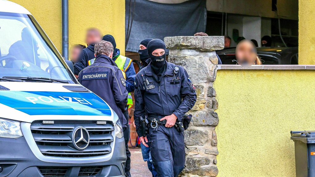 Bewaffnete Polizisten mit Sturmhaube stehen vor einem Haus vor dem ein Polizeiauto parkt