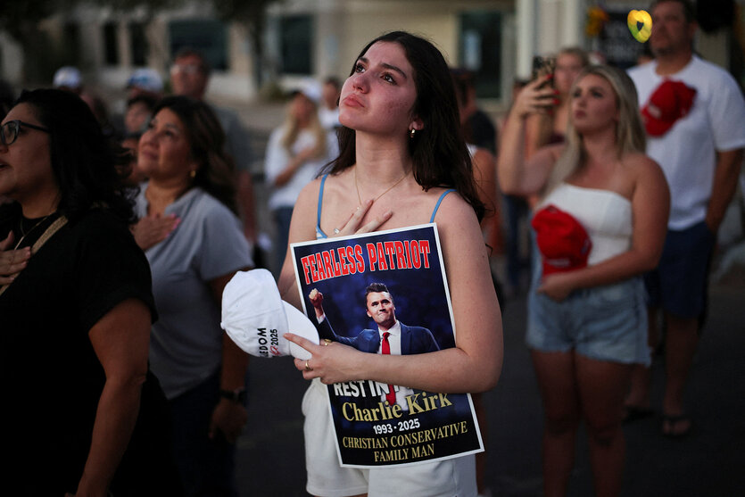 Eine Frau hält eine Kappe in den Händen und hat ein Poster mit einem Bild von Charlie Kirk unter ihren Arm geklemmt