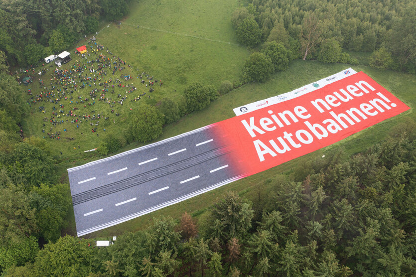 Ein riesiges Banner mit den Worten "Keine neuen Autobahnen" liegt auf einer Lichtung. Daneben stehen Menschen.