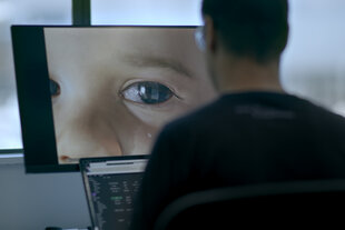 Ein Mann sitzt vor einem Bildschirm, der ein großes Auge mit einer Träne in einem Kindergesicht zeigt, während ein Laptop mit Code vor ihm offen ist