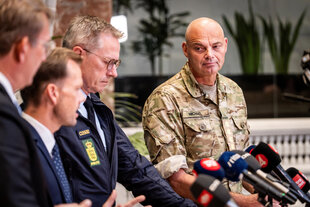 Drei Männer in Zivilkleidung (Anzug) und ein Mann in Militäruniform stehen vor den Mikrofonen der PResse mit ernstem Gesichtsausdruck