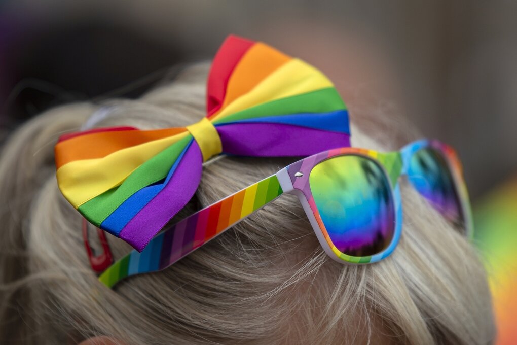 Jemand trägt eine Schleife in Regenbogenfarben im haar und hat eine regenbosenfarbige Sonnenbrille ins haar gesteckt