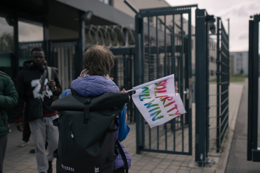Eine Mensch steht vor einem Zaungitter, in der Ahnd eine Fahne mit derr Aufschrift "Solidarity will win".