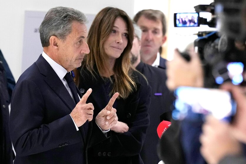 Nikolas Sarkozy steht mit erhobenen Zeigefingern vor den Kameras der Presse, neben ihm seine Frau Carla Bruni