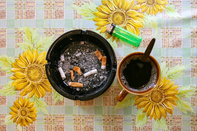 Ein voller Aschenbecher, eine Tasse mit schwarzem Kafee und ein grünes feuerzeug auf einer Wachstuchtischdecke mit Sonnenblumenmuster