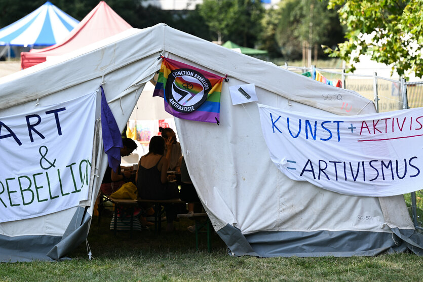 Teilnehmer des „System Change Camp“ für Klimagerechtigkeit sitzen im Grüneburgpark in einem Zelt, an dem Plakate mit der Aufschrift „Art & Rebellion“ (l) und „Kunst + Aktivismus = Artivismus“ angebracht sind. Zu den Mitveranstaltern des Treffens linker Gruppierungen zählen unter anderem die Klima-Aktivsten von Extinction Rebellion und das global..