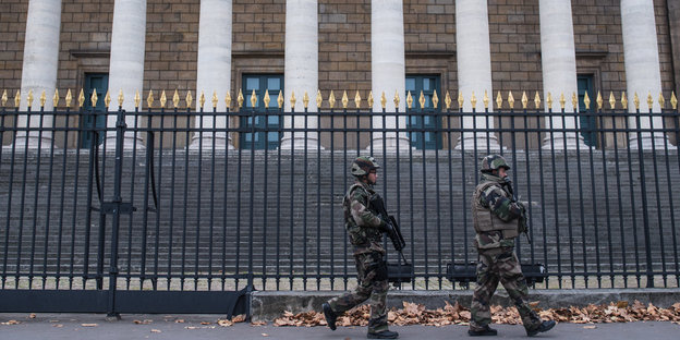 Zwei Männer mit Waffen patrouillen vor einem Gebäude.