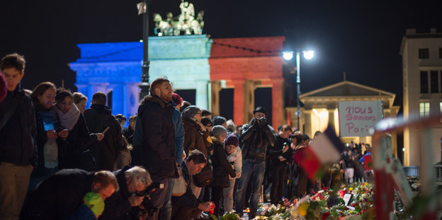 Menschen stehen an einer Streifen aus Blumen und Kerzen. Im Hintergund das Brandenburger Tor in blau, weiß, rot.