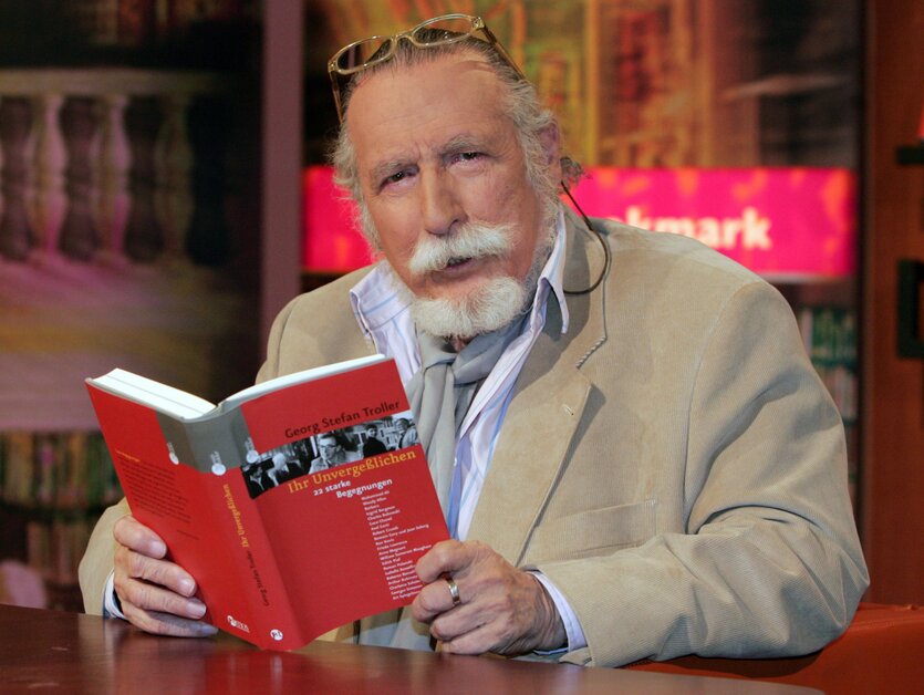 Troller hat einen schönen weißen Schnauzbart und hält ein rotes Buch