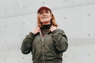Portrait von Caren Lay in olivgrünerBomberjacke und rosa Cap. Sie trägt eine Kette mit der Aufschrift Mietendeckel