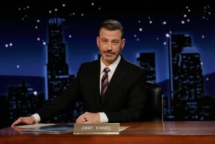 Jimmy Kimmel sitzt bei seiner Show an einem hölzernen Schreibtisch. Er hat eine Hand vor sich gelegt. die andere greif an den Rand des Tisches. Er trägt einen Anzug und Krawatte. Kimmel hat dunkle, kurze Haare.