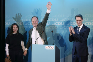 Eine Frau und zwei Männer hinter einem Rednerpult, einer der Männer winkt