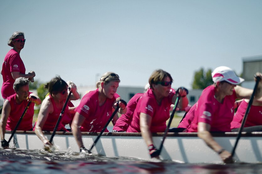 Sechs Frauen mit pinkfarbenen Trikots in einem Drachenboot. Fünf paddeln, die sechste steuert im Stehen.