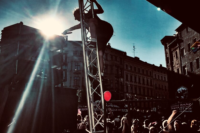 Ein Mann mit Mikrophon ist an den Seitenständern einer Bühne hochgekletter. Die Sonne knallt auf die Szene. Unten Publikum.