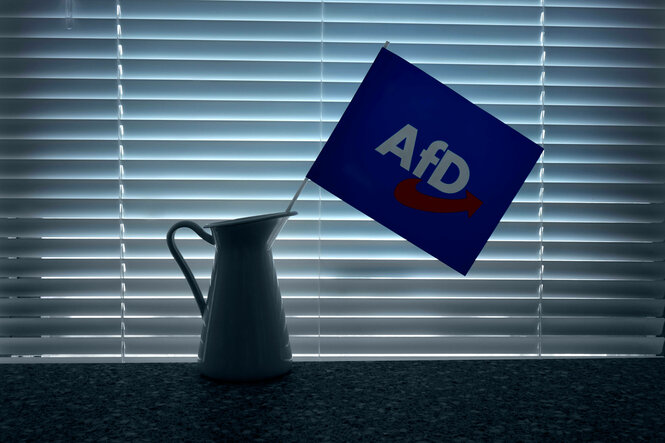 AfD-Fahne in einer Blechkanne vor Jalousie