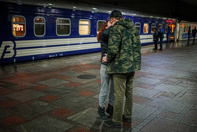 Ein Paar verabschiedet sich am Gleis mit einem Kuss. Die kleinere Person trägt eine helle Jeans, eine dunkle Jacke und hat lange, offene rote Haare. Ihre Hände liegen auf dem Nacken der größeren Person auf. Die größere steht mit dem Rücken zur Kamera, trägt eine Kappe und eine Jacke mit Tarnmuster und hält die kleinere Person an der Taille fest. Hinter dem Paar wartet ein ukrainischer Nachtzug.