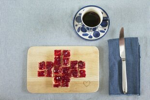 Auf hellblauer Tischdecke: Brettchen mit Marmeladentost in Kreuzform, Tasse Kaffe in Blümchenporzellan, Messer auf karierter Serviette,