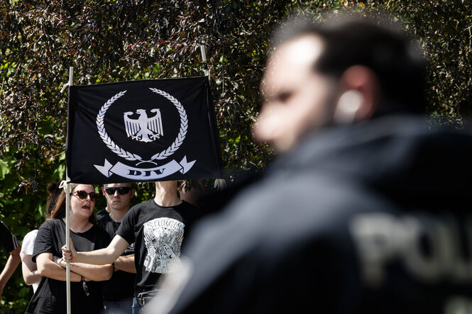DJV-Aufmarsch in Berlin: Ein Polizist im Vordergrund, hinten halten Menschen eine schwarze Fahne mit Adler und Aufdruck „DJV“