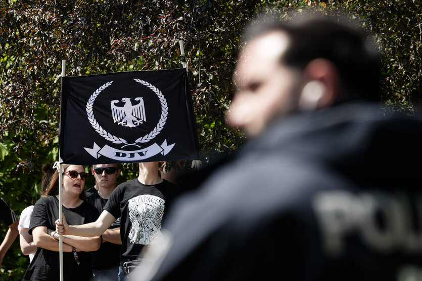 DJV-Aufmarsch in Berlin: Ein Polizist im Vordergrund, hinten halten Menschen eine schwarze Fahne mit Adler und Aufdruck „DJV“