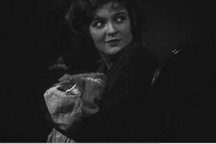 Aufnahme aus einem Schwarz-Weiß-Film. Die Schauspielerin Anna Sten sitz auf einem Stuhl und wendet den Blick über die Schulter. Sie blickt aufmerksam hinter sich.