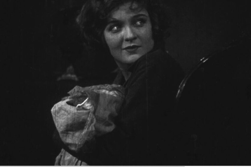 Aufnahme aus einem Schwarz-Weiß-Film. Die Schauspielerin Anna Sten sitz auf einem Stuhl und wendet den Blick über die Schulter. Sie blickt aufmerksam hinter sich.