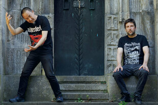 Die Musiker Yan Jun und Kai Fagaschinski stehen vor einem Metalltor, das mit Ornamenten verziert ist. Sie tragen Band-T-Shirts, auf dem T-Shirt von Kai Fagaschinski ist der Bandname "Saint Vitus" zu lesen. Yan Jun hebt die Hände, als halte er ein unsichtbares Objekt oder Energie in der Luft.
