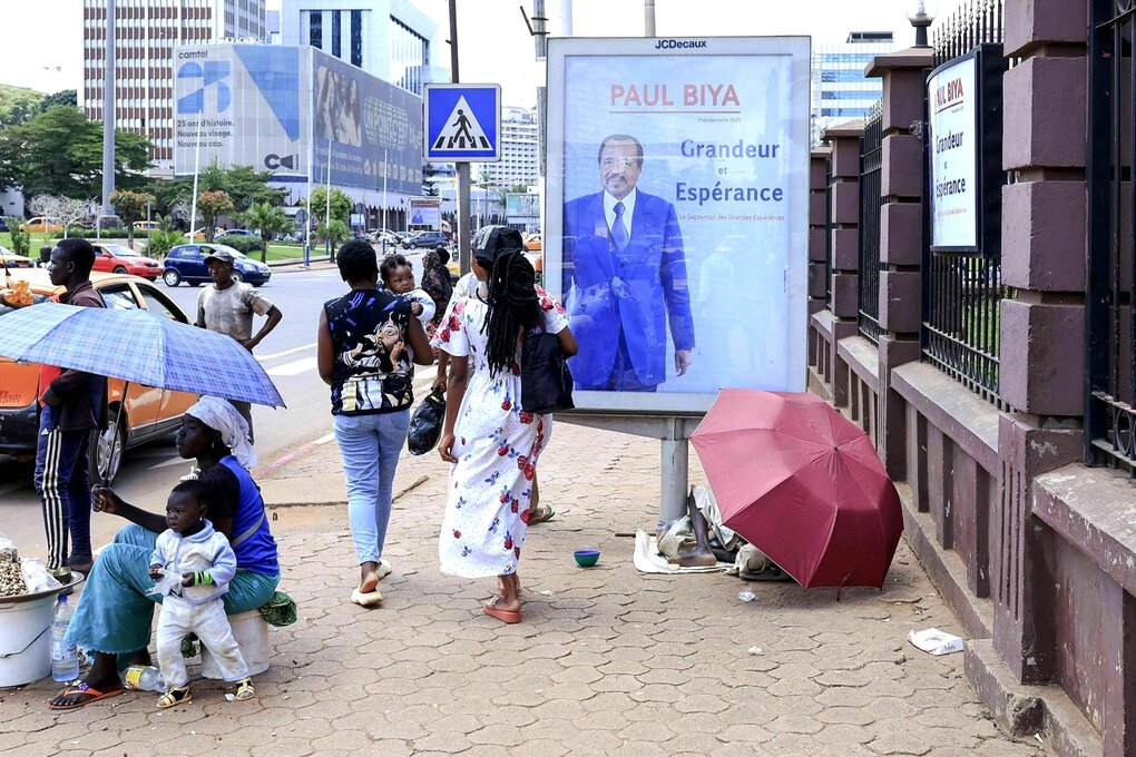 Auf dem Gehsteig ein Wahlplakat von Paul Biya - Fussgänger gehen vorbei, am Straßenrand verkauft eine Frau Lebensmittel, sie sitzt mit ihren Kindern unter einem Schirm
