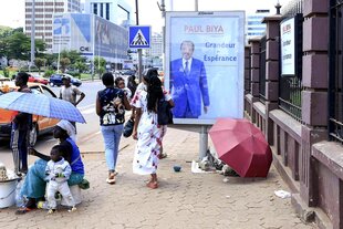 Auf dem Gehsteig ein Wahlplakat von Paul Biya - Fussgänger gehen vorbei, am Straßenrand verkauft eine Frau Lebensmittel, sie sitzt mit ihren Kindern unter einem Schirm