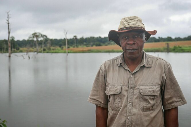 Julius Manjoh trägt einen Hut und ein helles Hemd und steht vor einer Wasserfläche