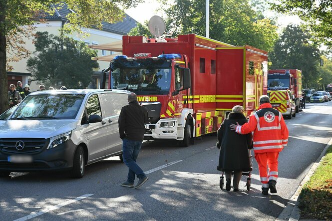 Ein Feuerwehrwagen steht in einer Straße in einer Wohnsiedlung. Im Vordergrund ein Rettungssanitäter und eine alte Person.