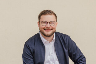 Ein junger Mann mit Brille und Bart lächelt in die Kamera vor einem beigen Hintergrund.