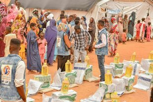 Frauen und Kinder stehen vor einem Zelt von Unicef - auf dem rötlichen Boden stehen aufgestapelte Grundnahrungsmittel