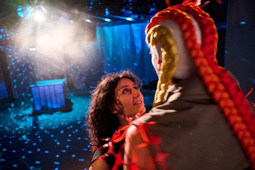 Theater-Szene: Eine Frau mit lockigem dunklem Haar schaut überrascht und emotional zu einer maskierten Figur mit weißem Gesicht, blonden Zöpfen, rotem Umhang und Federn empor, umgeben von blauen Spotlights, Nebel und einem podestartigen Bühnenaufbau.