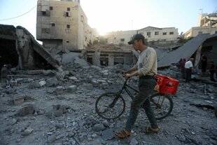 Ein Mann schiebt ein Fahrrad mit einem roten Plastikkorb über Trümmer und Schutt in einer zerstörten Straße in Gaza-Stadt. Im Hintergrund sind schwer beschädigte und eingestürzte Gebäude zu sehen. Mehrere Menschen durchsuchen die Trümmer. Die Szene spielt bei Tageslicht.