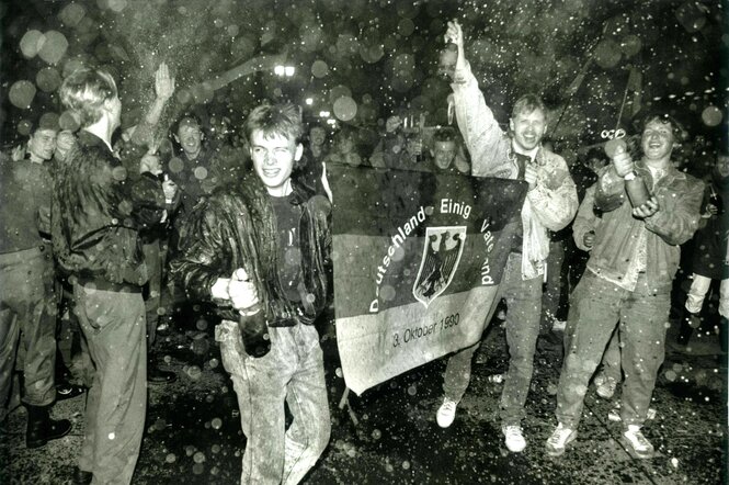 Junge Männer mit Sektflaschen und bester Laune hatlen eine Flagge in der Hand: Deutschland einig Vaterland 3. Oktober 1990