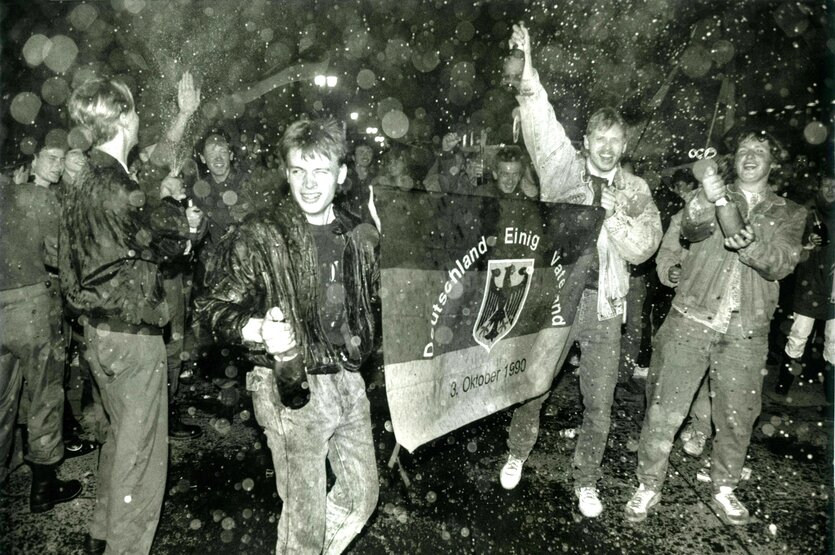 Junge Männer mit Sektflaschen und bester Laune hatlen eine Flagge in der Hand: Deutschland einig Vaterland 3. Oktober 1990
