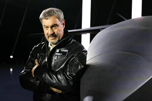 Markus Söder trägt eine schwarz-glänzende Lederjacke und lehnt mit verschränkten Armen an einem grauen Flugbjekt