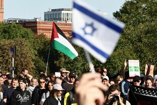 Eine israelische Flagge ist neben Menschen zu sehen, die demonstrieren. In der Mitte wird eine Palästinenserflagge hochgehalten, einige wenige tragen ein Pali-Tuch