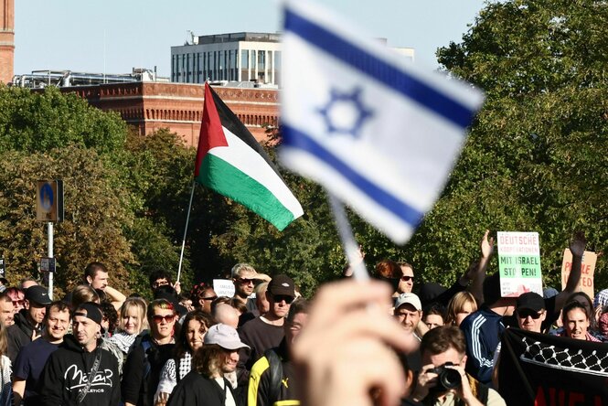Eine israelische Flagge ist neben Menschen zu sehen, die demonstrieren. In der Mitte wird eine Palästinenserflagge hochgehalten, einige wenige tragen ein Pali-Tuch