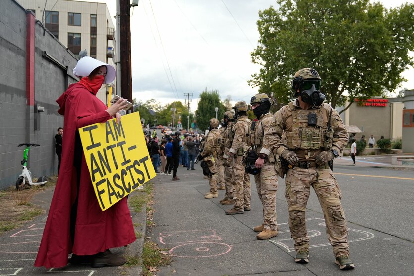 Frau mit einem Schid, auf dem steht: "I am antifascist". Daneben Uniformierte mit Helmen und Gasmasken