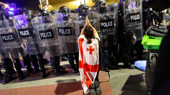 Eine Person mit einer georgischen Flagge umgehängt kniet vor einer Reihe von Polizisten