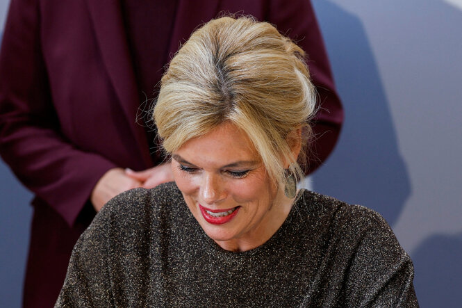 Eine blonde Frau mit aufgesteckten Haaren und rotem Lippenstift lächelt nach unten, während sie etwas unterschreibt. Sie trägt ein glitzerndes dunkelgraues Oberteil; im Hintergrund ist eine Person in einem bordeauxfarbenen Blazer zu sehen.