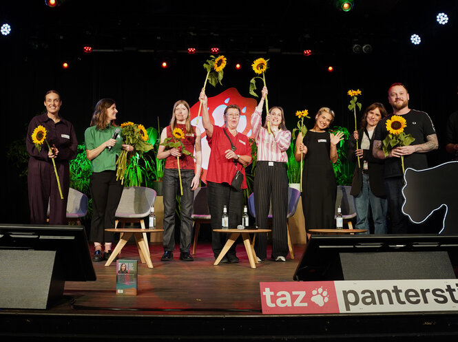 Die Bühne des Festsaals Kreuzberg während der Genoversammlung 2025. Im Hintergrund eine große taz-Tatze, davor das Genossenschaftsteam und Geschäftsführerin Aline Lüllmann, alle mit Sonnenblumen in der Hand.